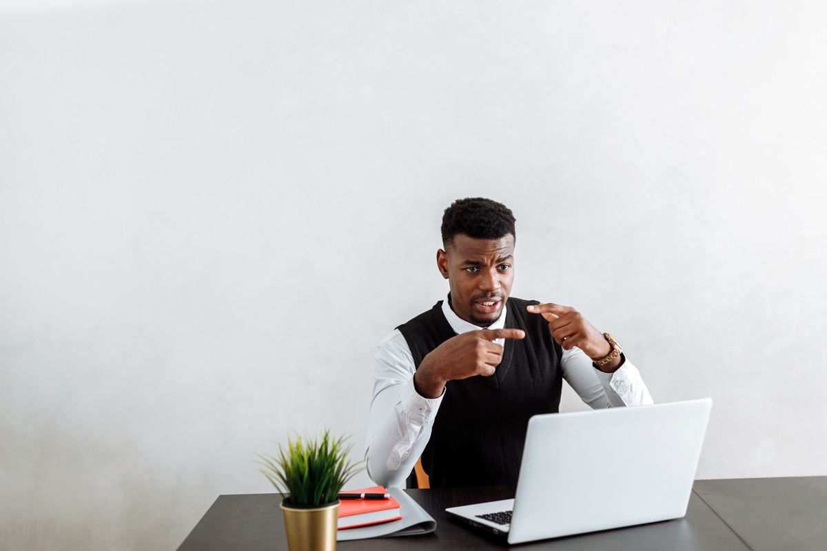 A man in a white shirt gesturing while explaining something on a laptop video call, like a candidate giving a too detailed behavioral interview answer