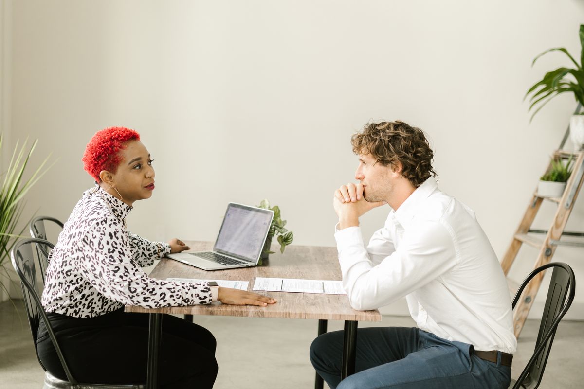 An interviewer and a candidate seated across a table in a bright office, the candidate listening with hands clasped as the interviewer answers
